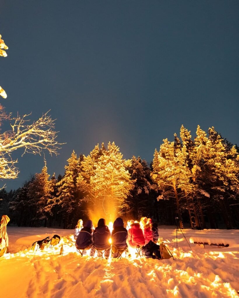 A group gathered around a campfire during a Northern Lights tour, sipping hot chocolate and grillng reindeer sausages. 