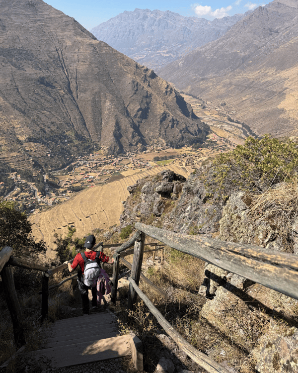 Hiker walking down stone steps with a wooden railing, with Pisac town and the Sacred Valley below